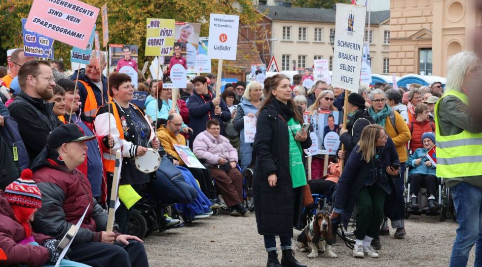 Viele Menschen nehmen an einer Demonstration teil, einige im Rollstuhl. Sie halten Plakate mit politischen Forderungen und Transparenten hoch.