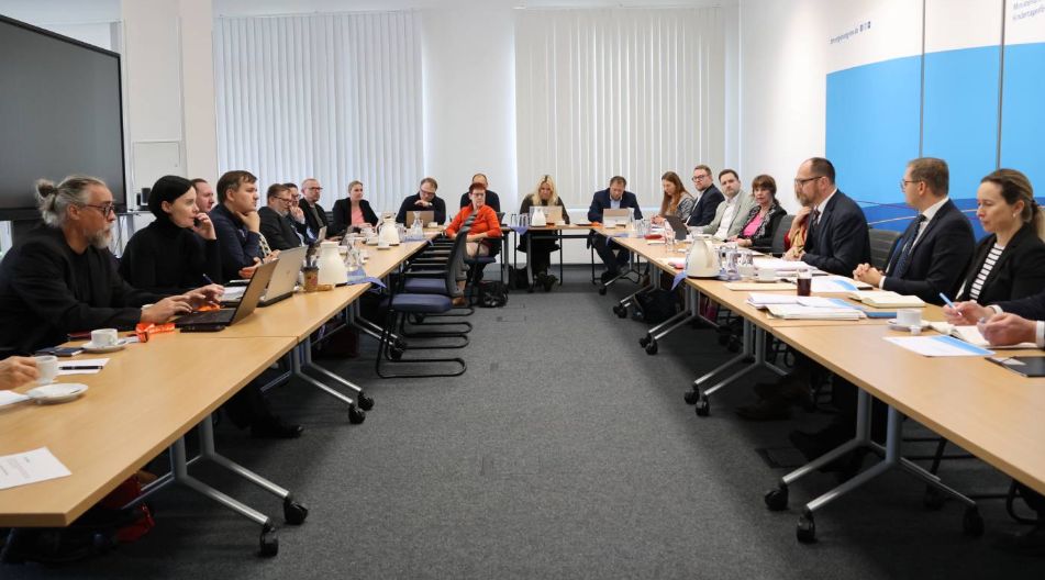 Mehrere Personen sitzen an zwei langen Tischen in einem Konferenzraum im Ministerium für Bildung und Kindertagesförderung und nehmen an einer Besprechung teil. Auf den Tischen liegen Unterlagen, Laptops und Kaffeekannen.