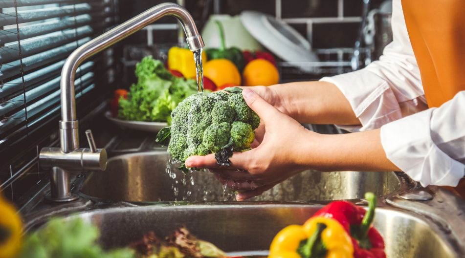 Eine Person wäscht frischen Brokkoli unter fließendem Wasser in der Küche, umgeben von buntem Gemüse wie Paprika und Salat.