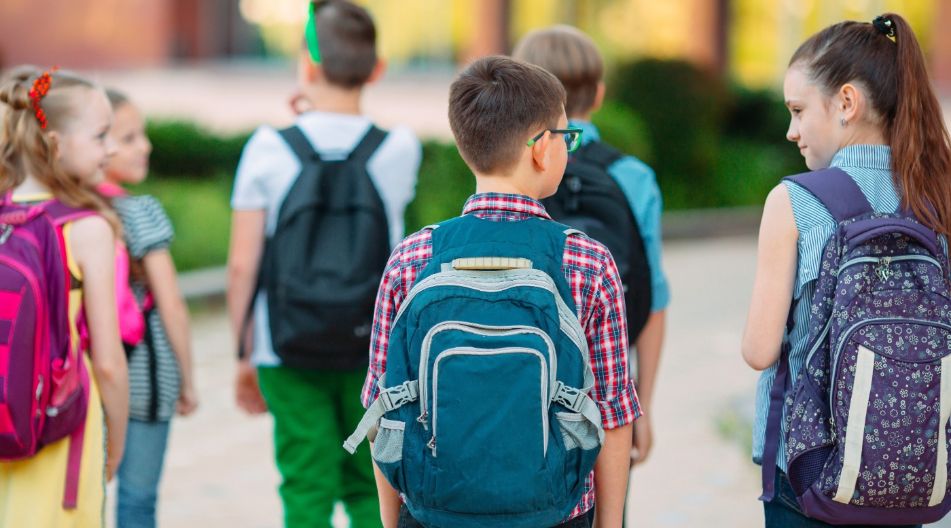 Eine Gruppe von Schulkindern mit Rucksäcken geht gemeinsam draußen, einige schauen sich an und unterhalten sich.
