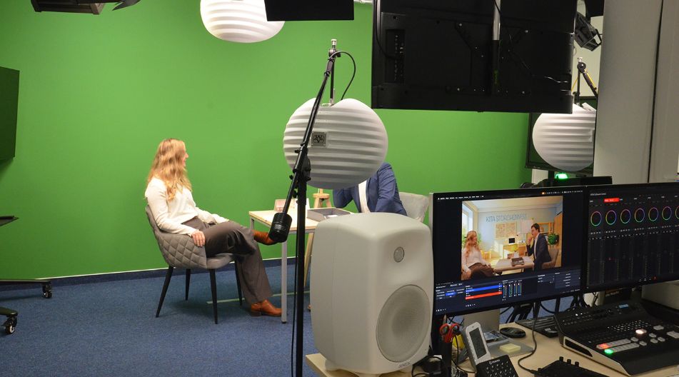 Zwei Personen sitzen sich in einem Studio mit grünem Hintergrund gegenüber, davor Monitor und technische Ausrüstung sichtbar Zwei Personen sitzen sich in einem Studio mit grünem Hintergrund gegenüber, davor Monitor und technische Ausrüstung sichtbar
