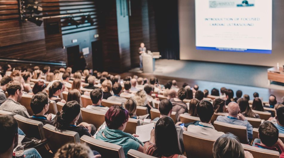 Ein großer Hörsaal mit vielen Zuhörern, die einer Präsentation auf einer Leinwand folgen. Eine Person steht am Rednerpult und hält einen Vortrag.