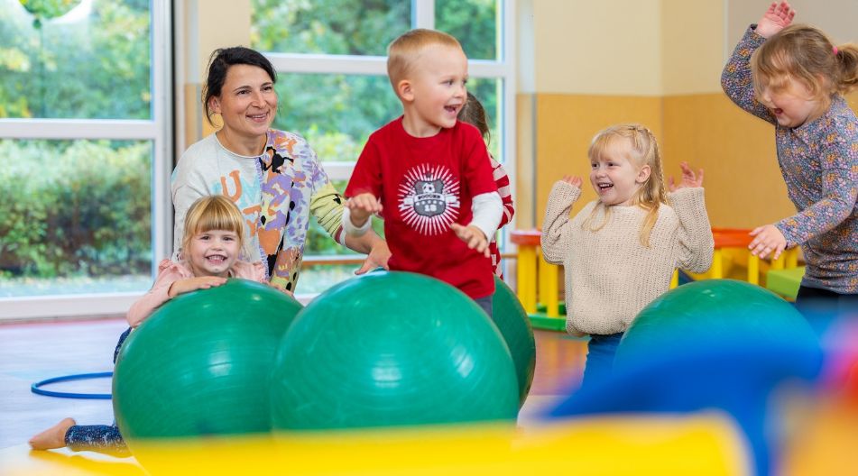 Kinder spielen lachend mit großen Gymnastikbällen, betreut von einer Erzieherin. Kinder spielen lachend mit großen Gymnastikbällen, betreut von einer Erzieherin.