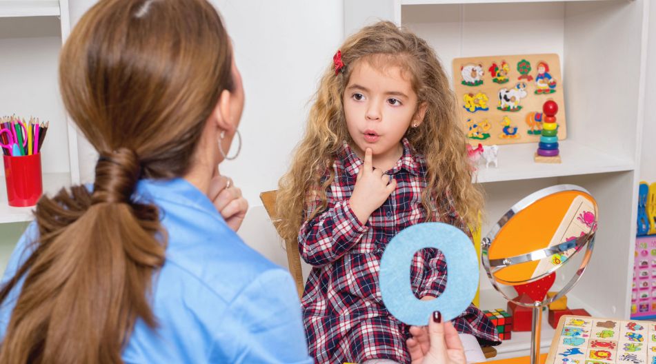 Eine Erzieherin sitzt mit einem jungen Mädchen in einem bunt gestalteten Lernraum. Das Mädchen trägt ein kariertes Kleid und formt mit den Lippen einen Laut, während die Frau einen blauen Buchstaben „O“ hält. Auf dem Tisch liegen Lernmaterialien, und ein Spiegel steht daneben.