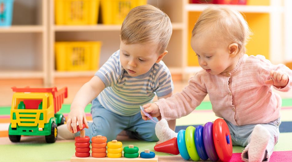 Zwei Kleinkinder spielen auf einer bunten Matte mit Lernspielzeug. Eines trägt ein gestreiftes Shirt und stapelt Ringe auf einem Holzstab, das andere trägt eine rosa Strickjacke und hält ein Plastikteil in der Hand. Im Hintergrund stehen Regale mit gelben Aufbewahrungskörben. Zwei Kleinkinder spielen auf einer bunten Matte mit Lernspielzeug. Eines trägt ein gestreiftes Shirt und stapelt Ringe auf einem Holzstab, das andere trägt eine rosa Strickjacke und hält ein Plastikteil in der Hand. Im Hintergrund stehen Regale mit gelben Aufbewahrungskörben.