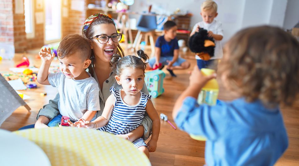 Ein heller und farbenfroher Kindergartenraum mit spielenden Kindern. Eine lachende Erzieherin sitzt mit zwei kleinen Kindern auf dem Boden, während andere Kinder um sie herum mit Spielzeug beschäftigt sind.