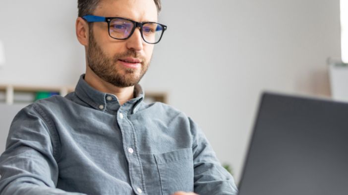 Ein Mann mit blauer Bildschirmarbeitsplatzbrille sitzt an einem Schreibtisch und arbeitet konzentriert an einem Laptop. Ein Mann mit blauer Bildschirmarbeitsplatzbrille sitzt an einem Schreibtisch und arbeitet konzentriert an einem Laptop.