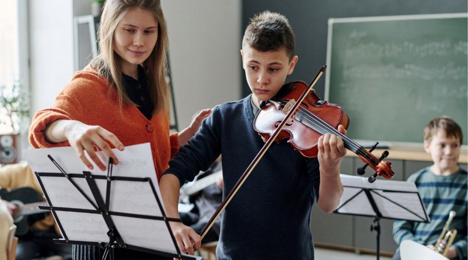 Eine Musiklehrerin hilft einem Jungen beim Geigenspiel, indem sie das Notenblatt richtet. Der Junge hält die Geige konzentriert, im Hintergrund sind weitere Kinder mit Musikinstrumenten in einem Klassenraum zu sehen.