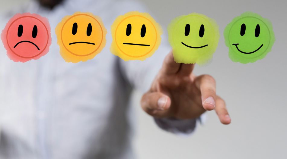 Eine Person im weißen Hemd zeigt auf eine Auswahl von fünf farbigen Smiley-Gesichtern, die von einem roten traurigen Gesicht bis zu einem dunkelgrünen sehr glücklichen Gesicht reichen, und wählt ein positives, grün lächelndes Gesicht aus.